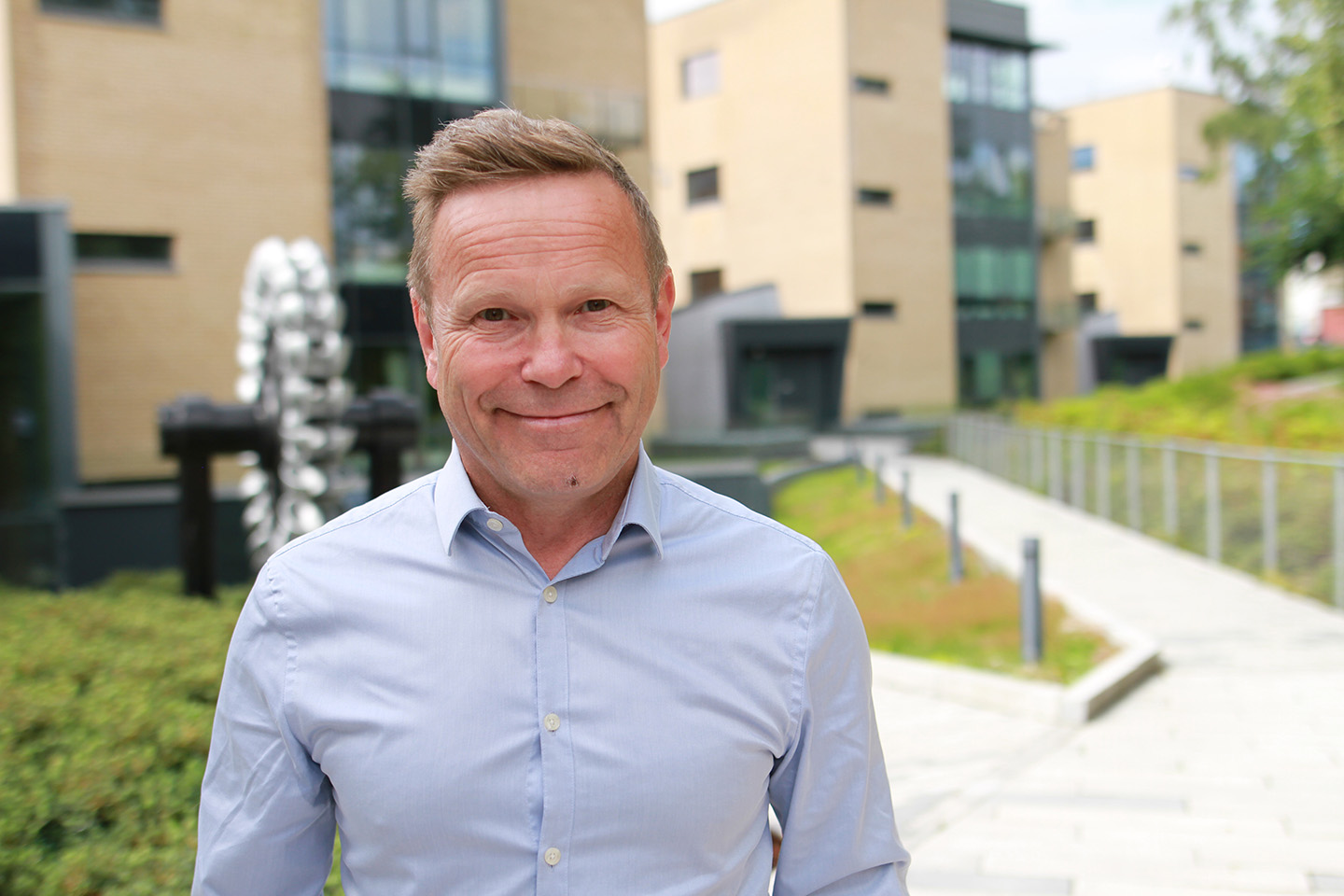 Mann med blondt hår står foran kontorbygg i blå skjorte og smiler til kamera.