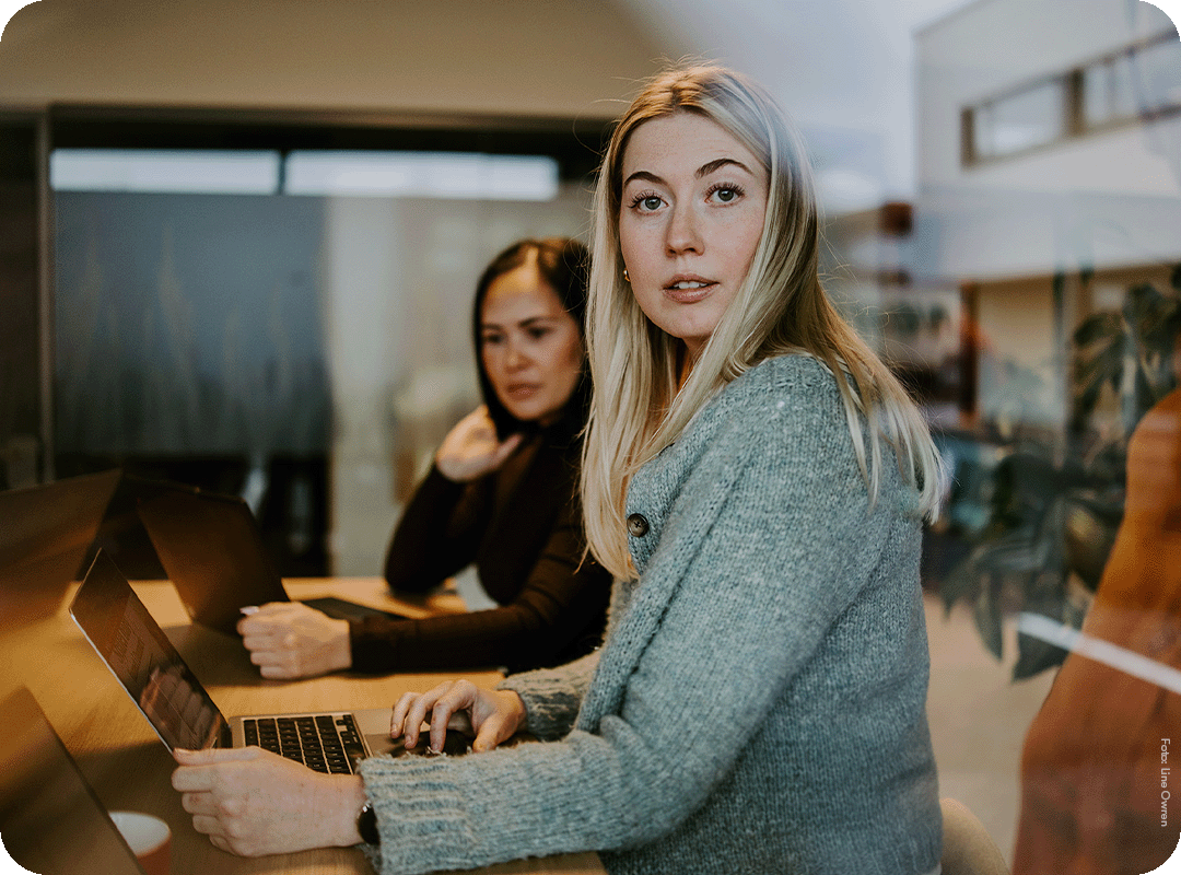 Kvinne ser mot kamera og sitter foran en laptop